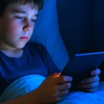 Niño acostado de madrugada frente a una tablet, iluminado por luz azul y con un reloj marcando la 1:15 a.m., símbolo del exceso de pantallas en la infancia.